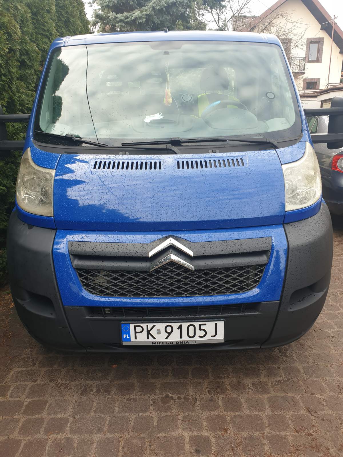 Przód niebieskiego busa marki Citroen z widoczną tablicą rejestracyjną PK 9105J, zaparkowanego na brukowanym podjeździe w pochmurny dzień.