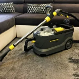 Szaro-żółty odkurzacz piorący Karcher na tle dywanu z krótkim włosiem i narożnika w odcieniach brązu z poduszkami w biało-czarne wzory.