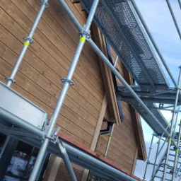 Fragment drewnianego domu w trakcie budowy, z widocznym rusztowaniem metalowym i elementami konstrukcyjnymi dachu.