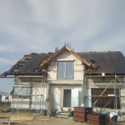 Dom w trakcie prac dekarskich: częściowo położona dachówka w kolorze grafitowym, widoczne rusztowania, palety z dachówką na ziemi, okno z PCV.