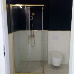 Nowoczesna łazienka z kabiną prysznicową w złotych ramach, biało-granatowe płytki, marmurowa podłoga i wisząca toaleta.