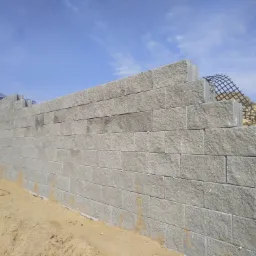 Wysoka ściana oporowa z szarych, betonowych bloków, wzmocniona siatką geosyntetyczną na szczycie, w tle błękitne niebo z chmurami.