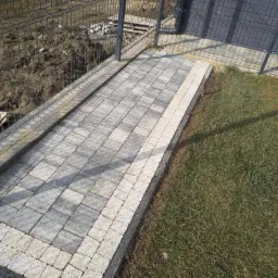 Świeżo ułożony chodnik z szarej kostki brukowej, obramowany jasną kostką, biegnący wzdłuż metalowego ogrodzenia na granicy trawnika i ziemi z gruzem.