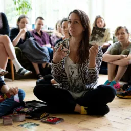 Kobieta w bluzie w zebrę prezentuje kartę z gry Awkward Family Photos grupie os&oacute;b siedzących na podłodze i kanapie podczas warsztat&oacute;w.