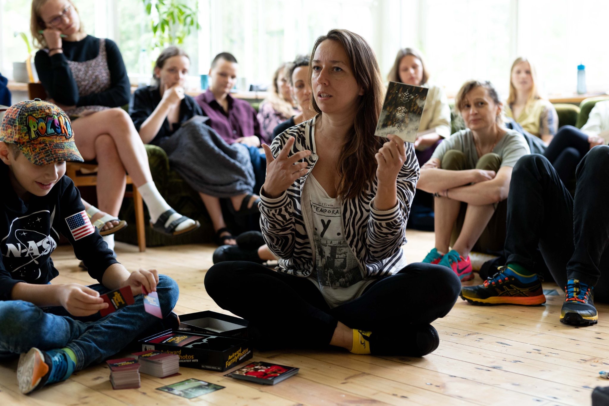 Kobieta w bluzie w zebrę prezentuje kartę z gry Awkward Family Photos grupie osób siedzących na podłodze i kanapie podczas warsztatów.