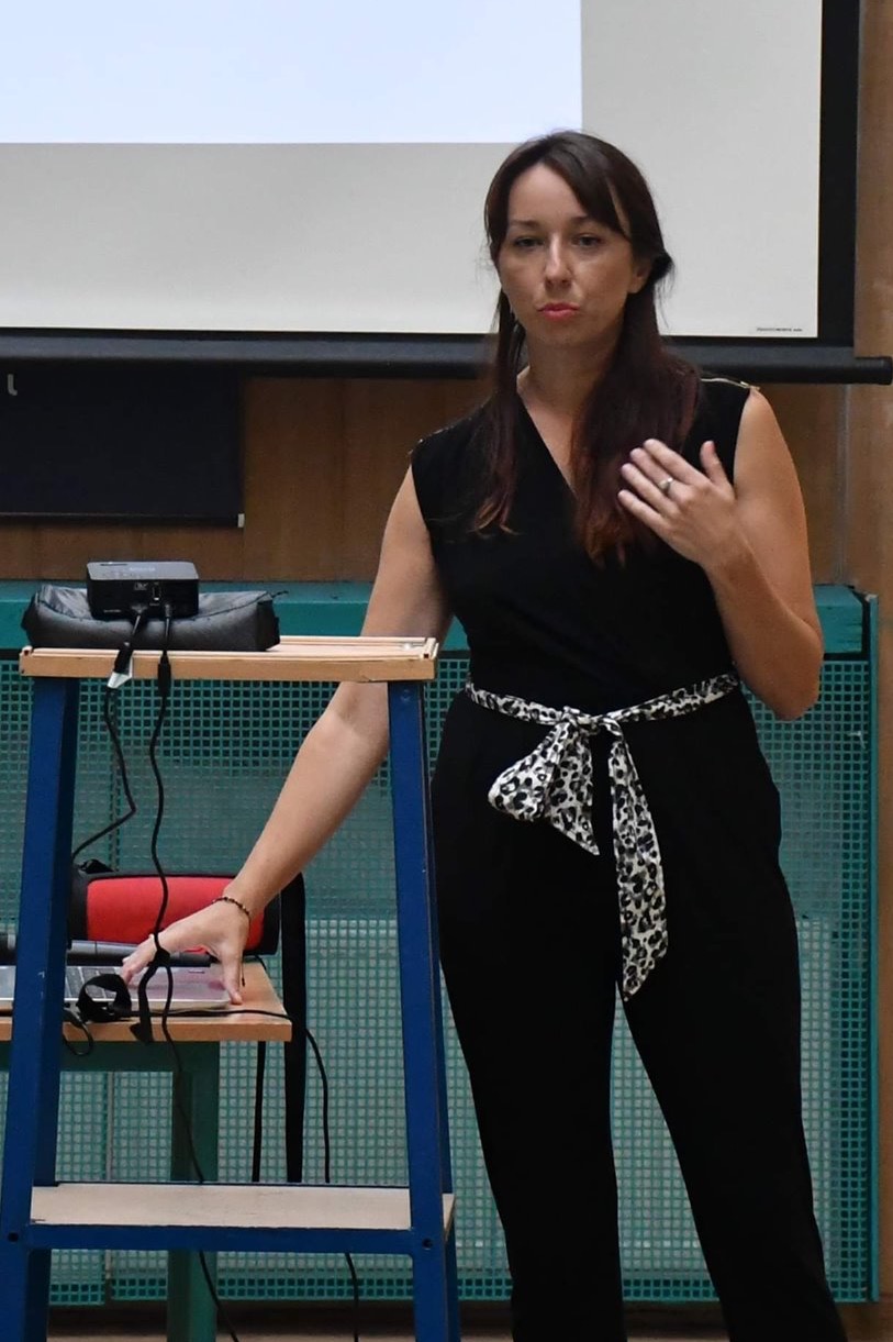 Kobieta w czarnym kombinezonie z szarfą w panterkę, gestykuluje podczas prezentacji. Na pierwszym planie stolik z laptopem i projektorem, w tle ekran projekcyjny.