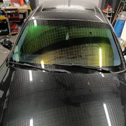 Ciemna szyba czołowa auta z delikatną zieloną poświatą, widok z zewnątrz, na pierwszym planie wycieraczki. Refleksy światła na karoserii.