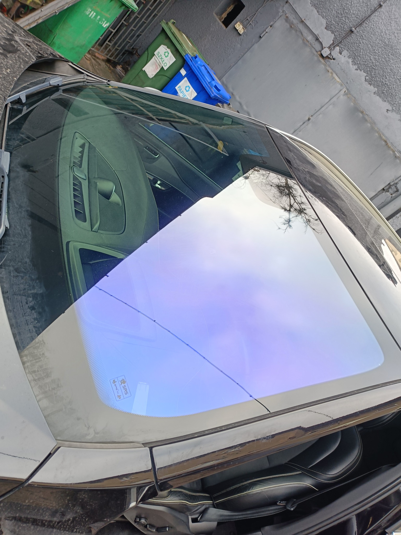 Auto z przyciemnioną szybą przednią, widoczne odbicie drzewa. Szyba z delikatnym niebieskim odcieniem. Ujęcie z góry, z boku auta. Na zewnątrz śmietniki.