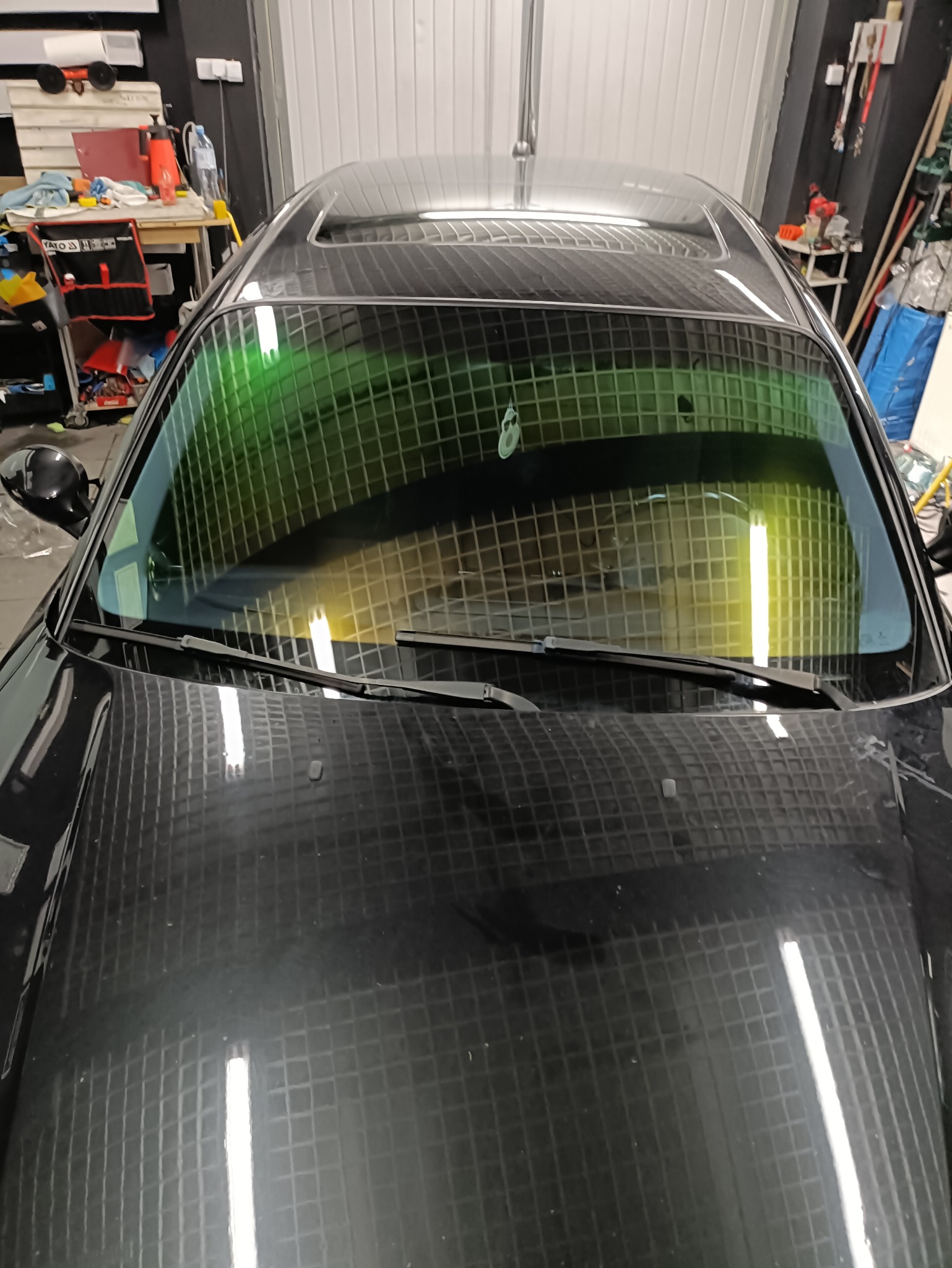 Ciemna szyba czołowa auta z delikatną zieloną poświatą, widok z zewnątrz, na pierwszym planie wycieraczki. Refleksy światła na karoserii.