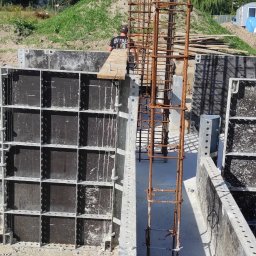 Feniiks Anna Cuber - Zbrojenie pionowe w szalunku fundamentowym, widoczna praca zbrojarza w tle, teren budowy z hałdą ziemi.