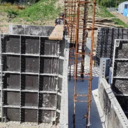 Zbrojenie pionowe w szalunku fundamentowym, widoczna praca zbrojarza w tle, teren budowy z hałdą ziemi.