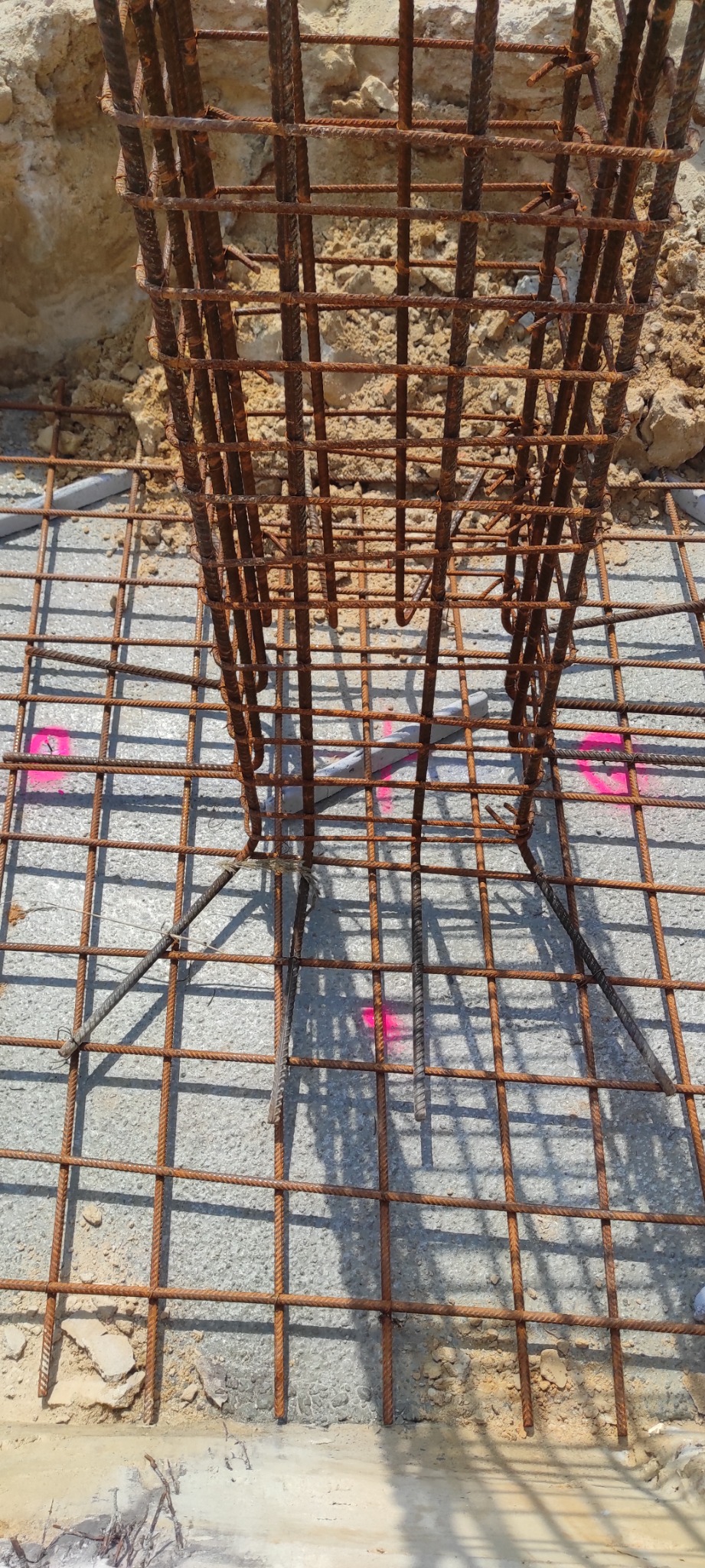 Siatka zbrojeniowa fundamentu z pionowymi prętami, widoczne różowe oznaczenia na betonie i fragment wykopu z ziemią w tle.