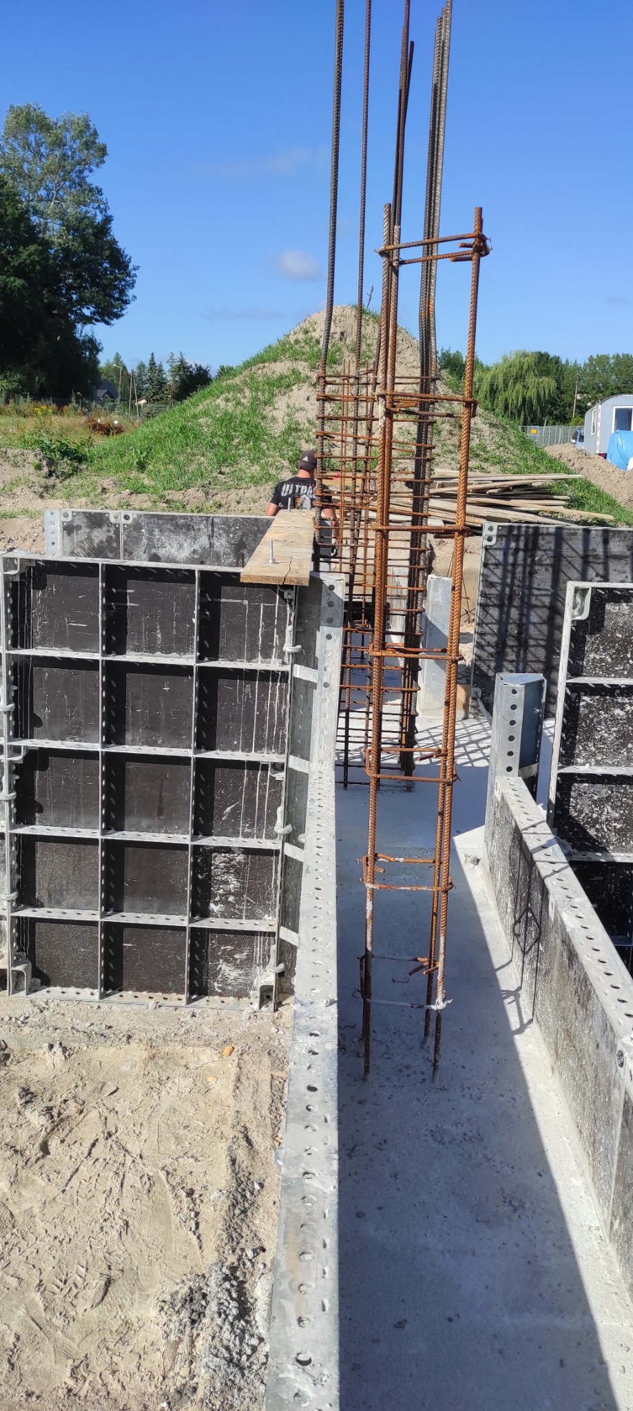Zbrojenie pionowe w szalunku fundamentowym, widoczna praca zbrojarza w tle, teren budowy z hałdą ziemi.