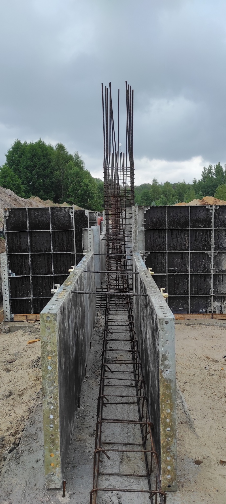 Szare betonowe szalunki z wystającymi prętami zbrojeniowymi, widok z góry na placu budowy w pochmurny dzień.