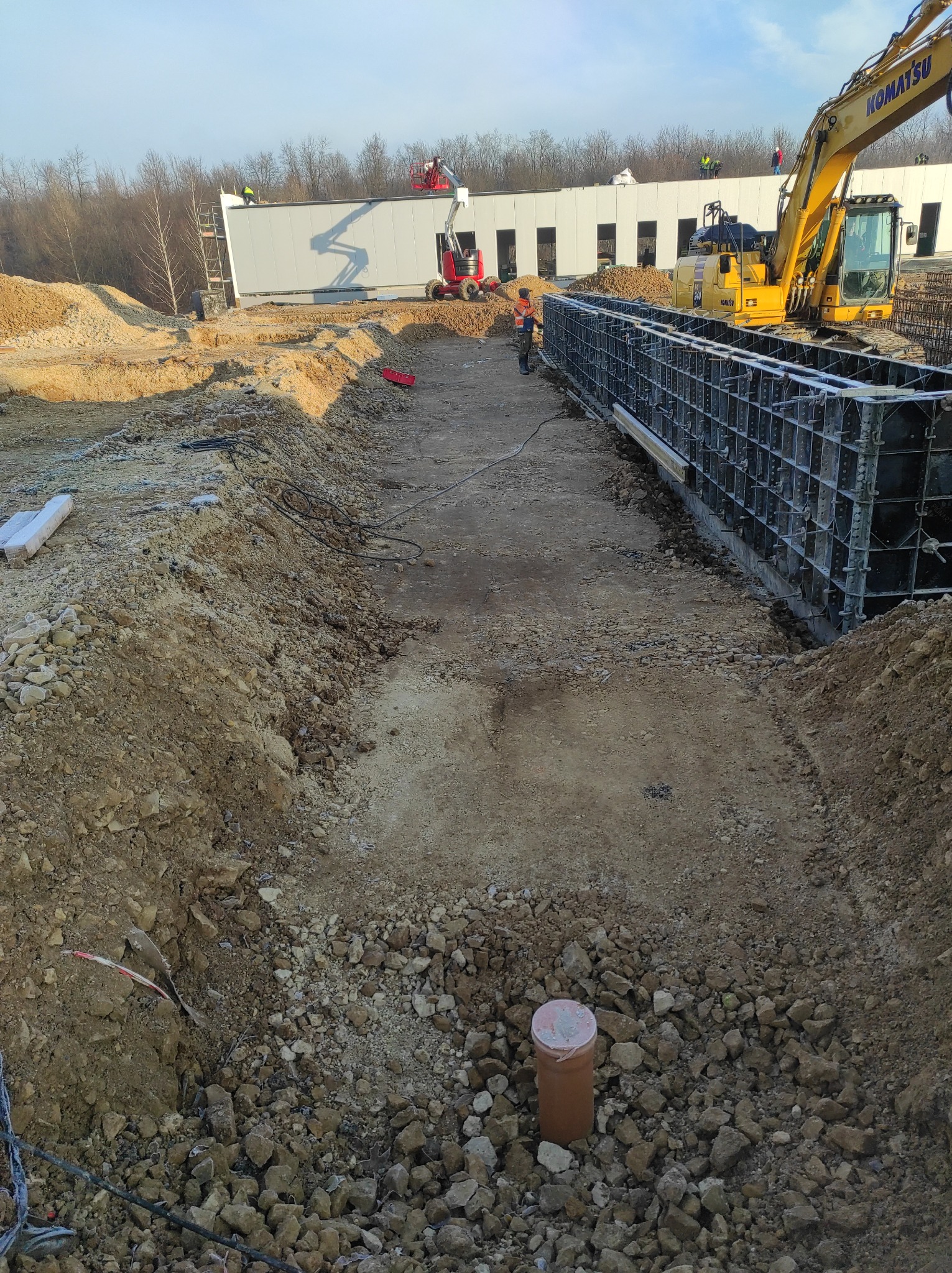 Przygotowanie wykopu pod fundamenty z szalunkami systemowymi, widoczna koparka Komatsu, pracownicy budowlani i budynek w budowie w tle.