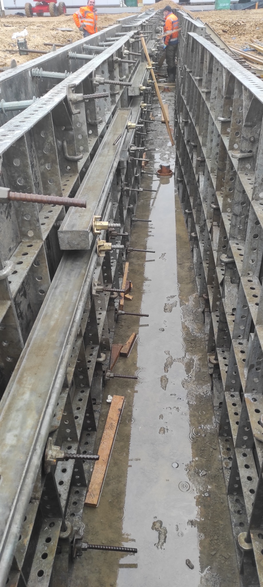 Metalowe szalunki zalane deszczem, widoczne pręty zbrojeniowe i pracownicy w odblaskowych ubraniach w tle.