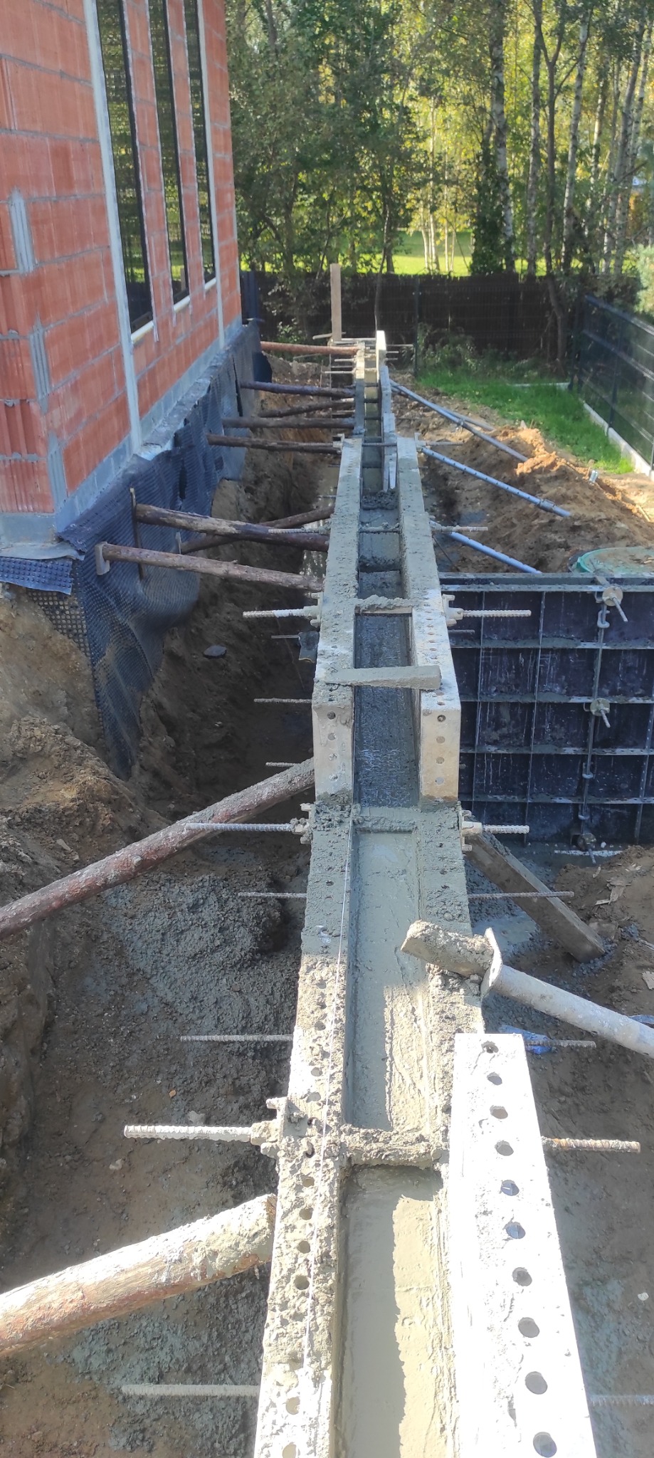 Wykop fundamentów z deskowaniem i zbrojeniem zalewanym betonem, widoczny fragment ściany budynku z czerwonej cegły.