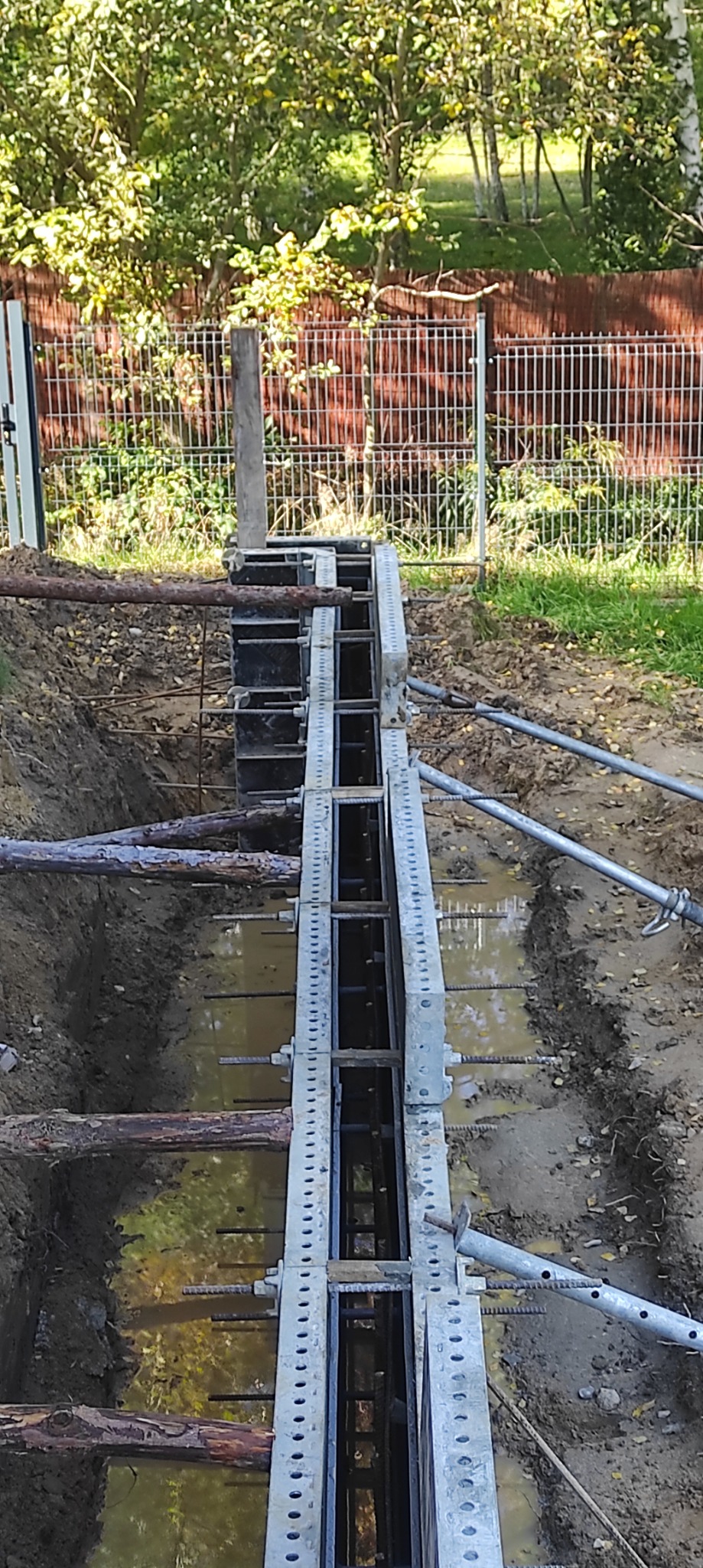 Szczegółowe ujęcie szalunku zbrojeniowego w wykopie wypełnionym wodą, zabezpieczonego drewnianymi belkami, z widocznym ogrodzeniem w tle.