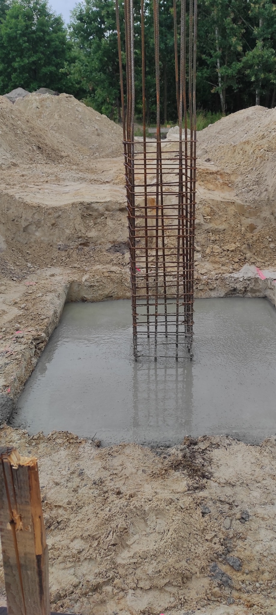 Kwadratowy fundament zalany mokrym betonem, wystająca zbrojona kolumna, widoczne wykopane ściany wykopu i hałdy ziemi w tle.