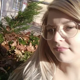 Portret kobiety w okularach na tle budynku z metalową barierką, iglakami i opadłymi liśćmi.