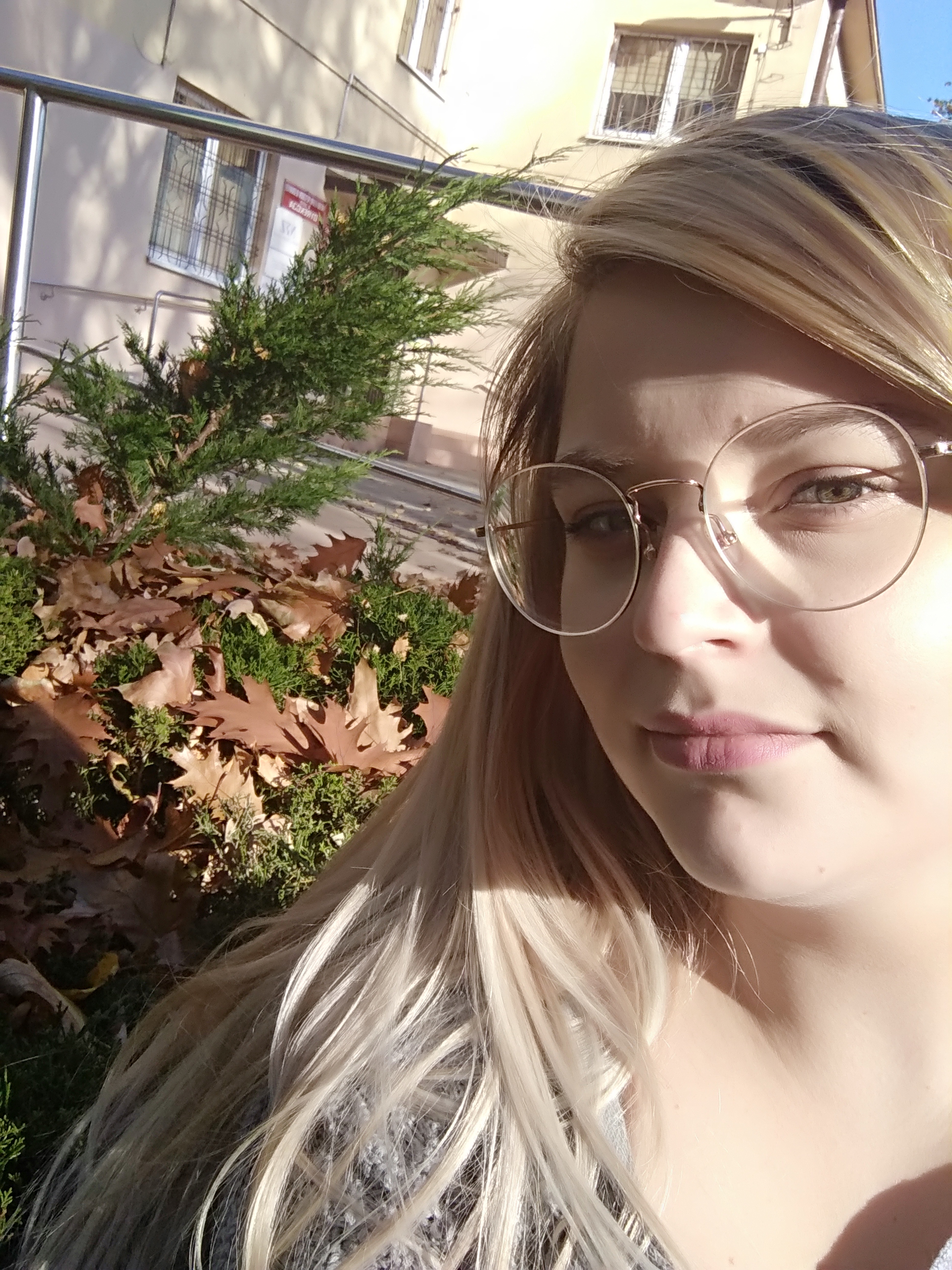 Portret kobiety w okularach na tle budynku z metalową barierką, iglakami i opadłymi liśćmi.