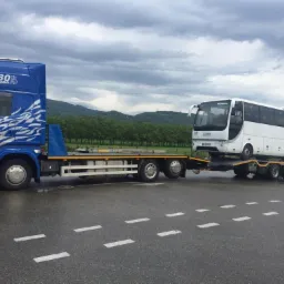 Niebieska laweta Scania R480 z białym autobusem Perraudo na platformie, transportowana po mokrym asfalcie na tle zalesionych wzg&oacute;rz pod pochmurnym niebem.