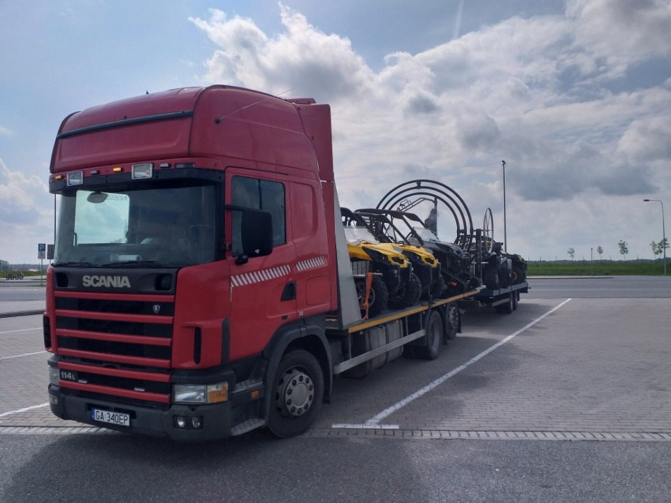 Czerwona ciężarówka Scania z naczepą przewożąca kilka żółtych i czarnych pojazdów typu buggy na parkingu z kostki brukowej, pochmurne niebo w tle.