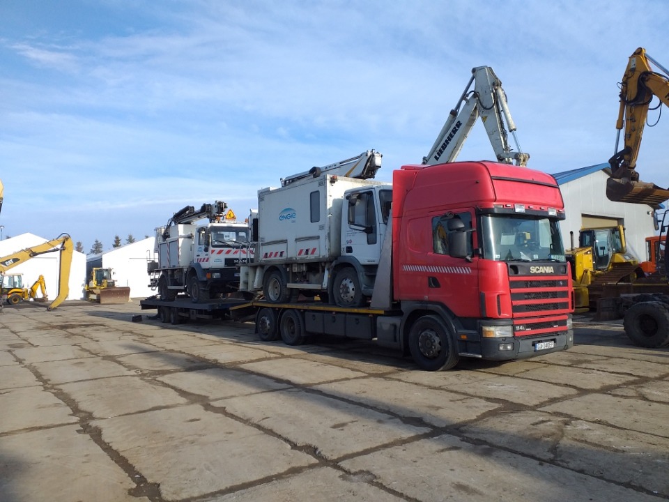 Czerwona ciężarówka Scania przewozi na lawecie dwa białe samochody serwisowe z dźwigami i logo ENGIE, na placu budowy z koparkami w tle.