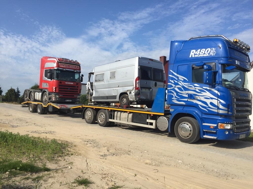 Dwa samochody ciężarowe typu laweta, jeden czerwony i jeden niebieski, przewożą pojazd typu kamper na platformie, na tle błękitnego nieba z chmurami.