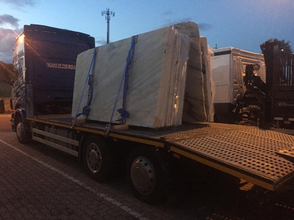 Ciężarówka z platformą załadowaną dużymi płytami marmuru zabezpieczonymi niebieskimi pasami transportowymi, widoczny napis 'KING OF THE ROAD' na kabinie.
