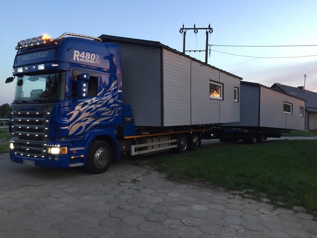 Niebieska ciężarówka Scania R480 z napisem 'King of the Road' przewozi modułowy dom o szarych ścianach z oknami, stojąc na betonowej nawierzchni przy zielonym trawniku.