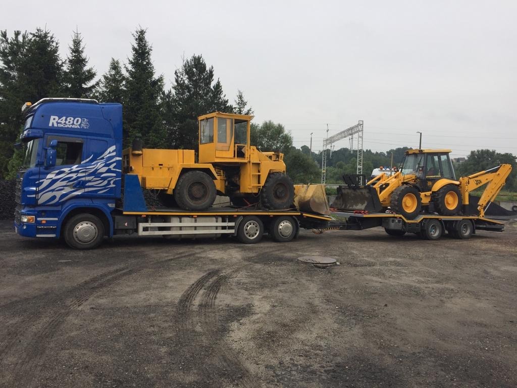 Niebieska ciężarówka Scania R480 z białymi płomieniami na kabinie transportuje na niskopodwoziowej lawecie dwie żółte maszyny budowlane: ładowarkę kołową i koparko-ładowarkę JCB.