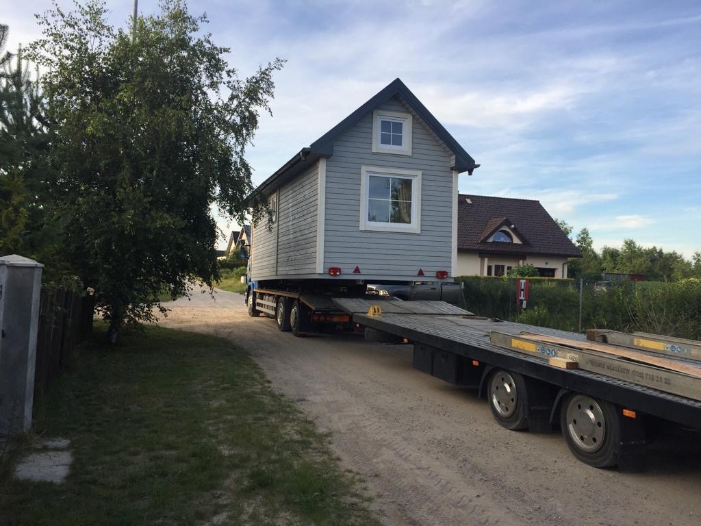 Przewóz modułowego domu w stylu skandynawskim na niskopodwoziowej naczepie transportowej po gruntowej drodze, widok z boku, w tle zabudowania jednorodzinne.