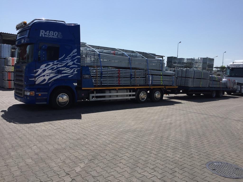Niebieska ciężarówka Scania R480 z naczepą przewożąca zabezpieczone metalowe rusztowania na placu wyłożonym kostką brukową, w tle składowane elementy rusztowań.