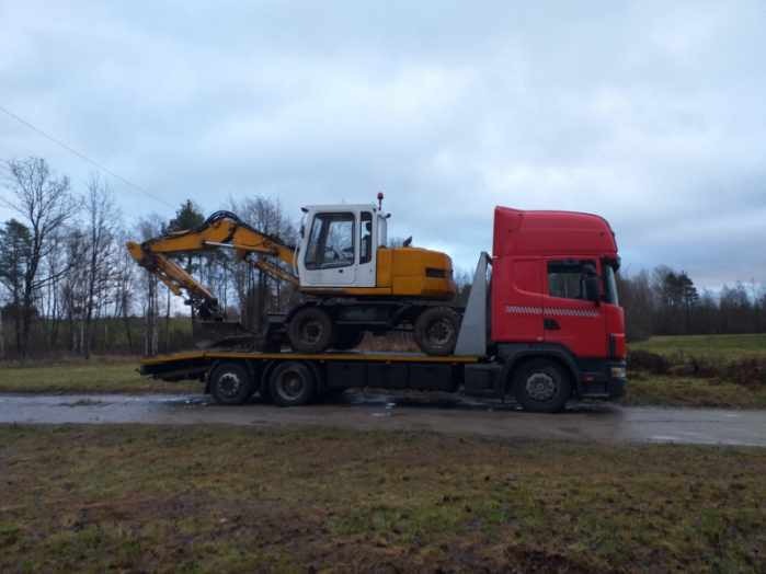 Czerwona ciężarówka Scania z żółto-białą koparką na niskopodwoziowej lawecie, transport po asfaltowej drodze na tle pola i drzew pod pochmurnym niebem.