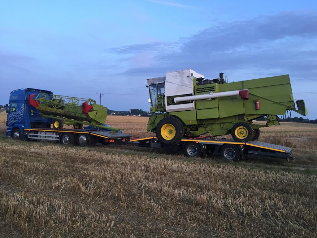 Niebieska ciężarówka przewozi na niskopodwoziowej lawecie kombajn zbożowy w kolorze zielonym i jego heder po polu ze ścierniskiem, widoczne zachodzące słońce.