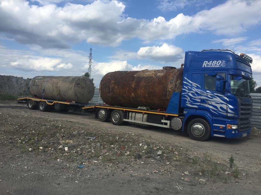 Niebieski ciągnik siodłowy Scania R480 z naczepą przewozi dwa duże, skorodowane zbiorniki na tle pochmurnego nieba i przemysłowego krajobrazu.