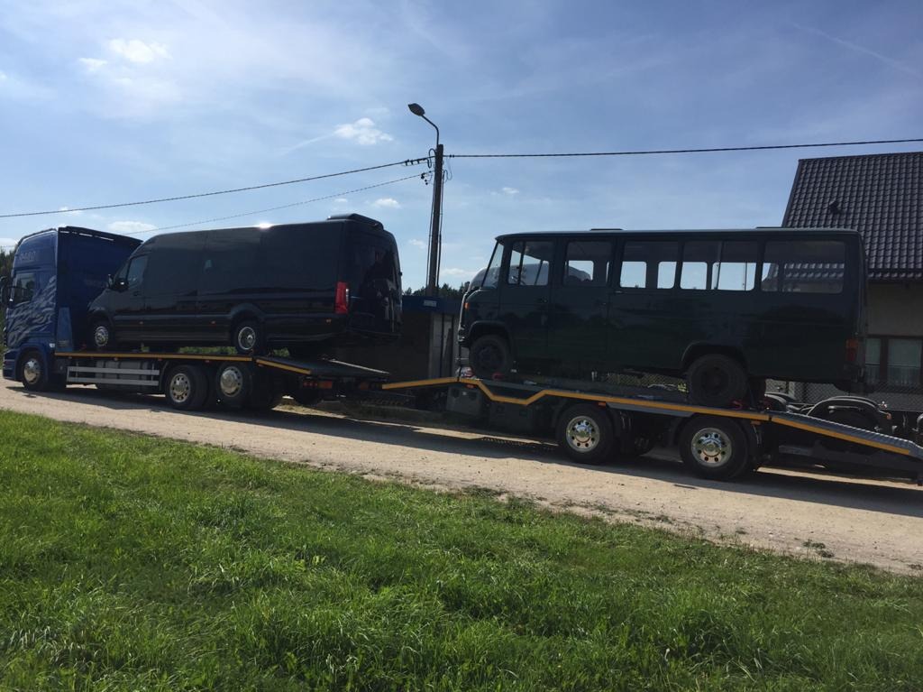 Dwa busy, czarny i ciemnozielony, przewożone na lawecie w Gdyni. Na pierwszym planie trawa, w tle dom z ciemnym dachem i słup elektryczny.