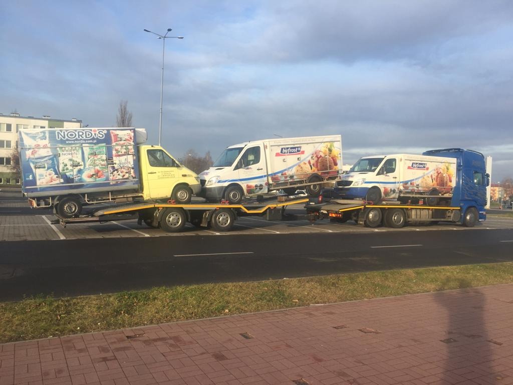 Dwa zestawy lawet transportują trzy samochody dostawcze z reklamami firm Nordis i Byfrost na parkingu.