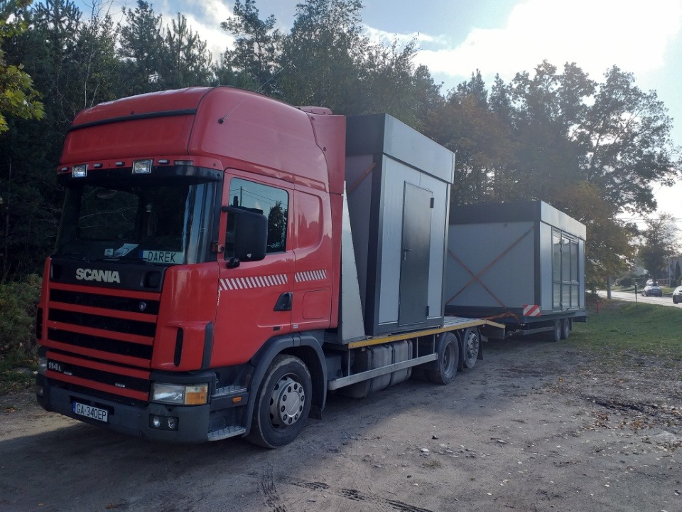 Czerwona ciężarówka Scania 114L z naczepą przewożąca dwie szare, modułowe konstrukcje kontenerowe z drzwiami i oknami, na tle lasu i błękitnego nieba.