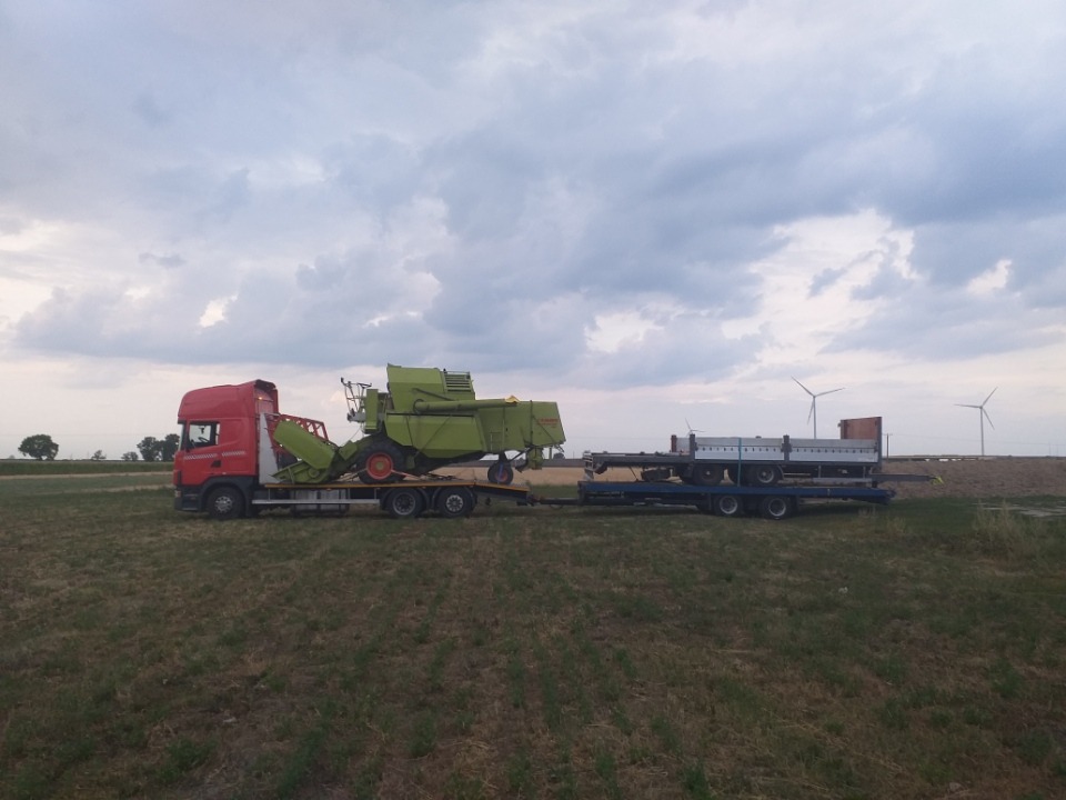 Czerwona ciężarówka z naczepą przewozi zielony kombajn rolniczy na tle pochmurnego nieba i turbin wiatrowych.
