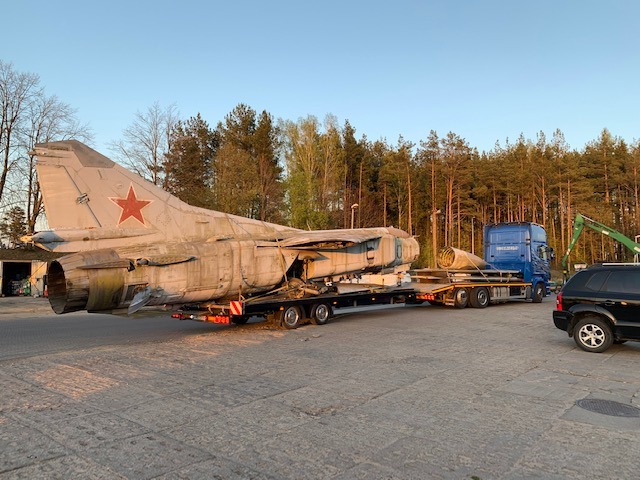 Transport uszkodzonego, radzieckiego samolotu myśliwskiego z czerwoną gwiazdą na ogonie na niskopodwoziowej platformie w Gdyni, z widocznym niebieskim ciągnikiem siodłowym i fragmentem lasu w tle.