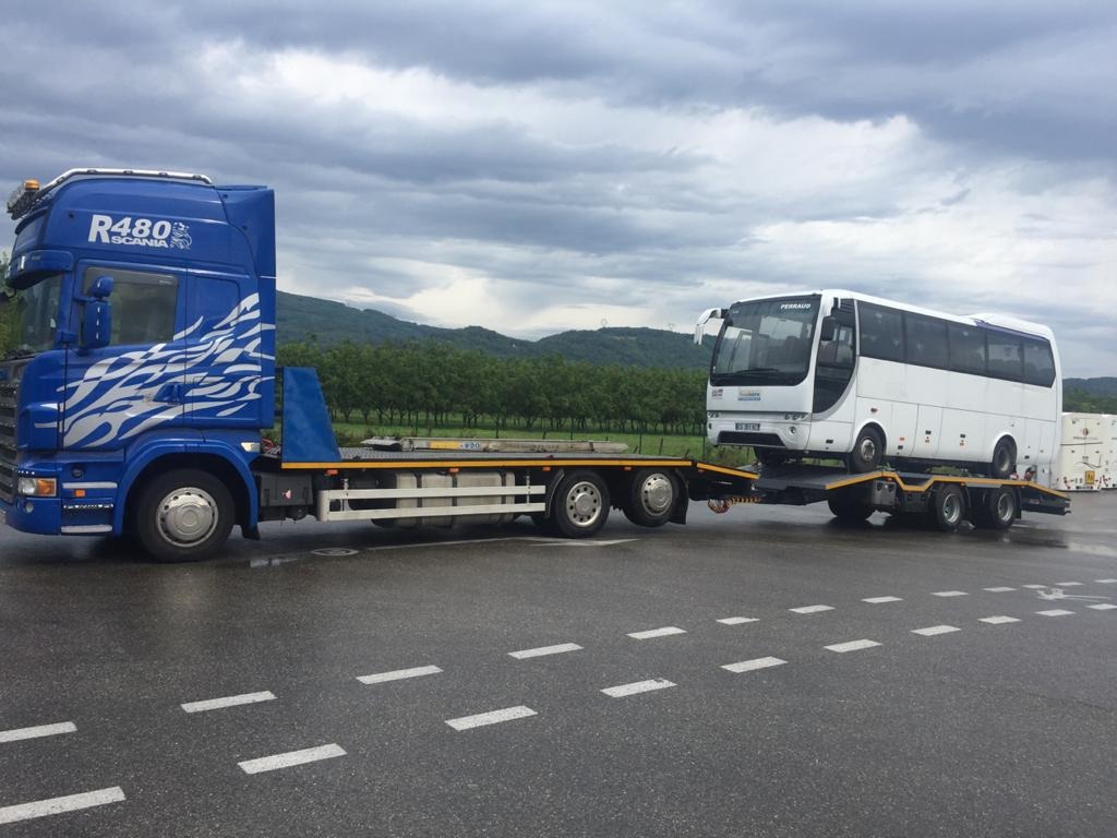 Niebieska laweta Scania R480 z białym autobusem Perraudo na platformie, transportowana po mokrym asfalcie na tle zalesionych wzgórz pod pochmurnym niebem.
