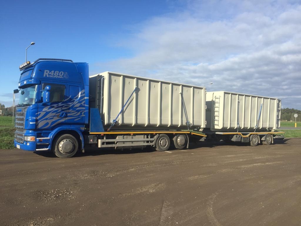 Niebieska ciężarówka Scania R480 z przyczepą przewożącą dwa duże, puste kontenery na tle pochmurnego nieba i zielonego krajobrazu.