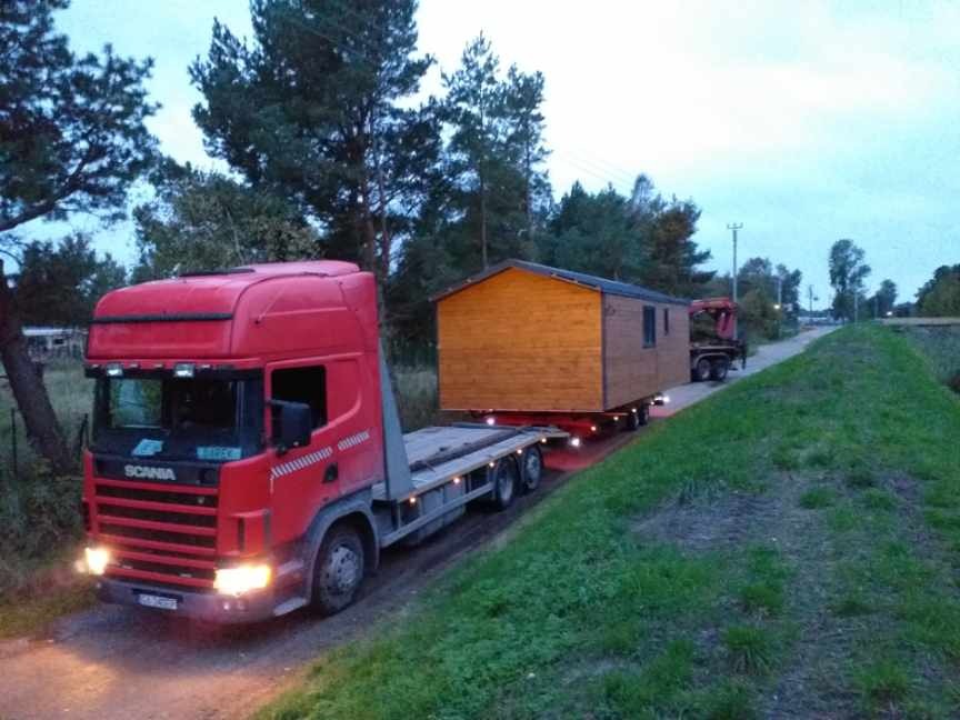 Czerwony ciągnik siodłowy Scania przewozi na niskopodwoziowej platformie drewniany modułowy dom oświetlony światłami pozycyjnymi, widok z boku na tle zieleni i drzew.