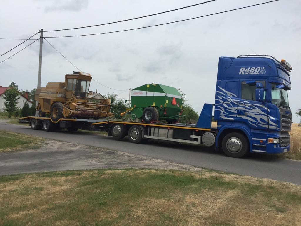 Niebieska ciężarówka Scania R480 z grafiką płomieni transportuje na niskopodwoziowej lawecie kombajn zbożowy SAMPO 500 i zieloną prasę rolującą po asfaltowej drodze w słoneczny dzień.