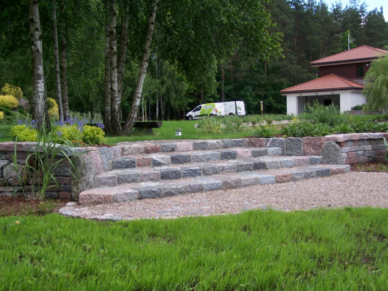 Granitowe stopnie prowadzące do ogrodu, z elementami muru oporowego, na tle zielonego trawnika i drzew, z widocznym samochodem dostawczym w oddali.
