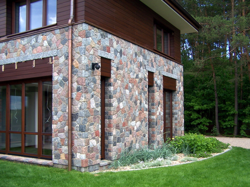 Fragment domu z elewacją z nieregularnych kamieni w odcieniach szarości, czerwieni i brązu, z ciemnobrązową drewnianą stolarką okienną i obudową, na tle zielonego trawnika i lasu iglastego.