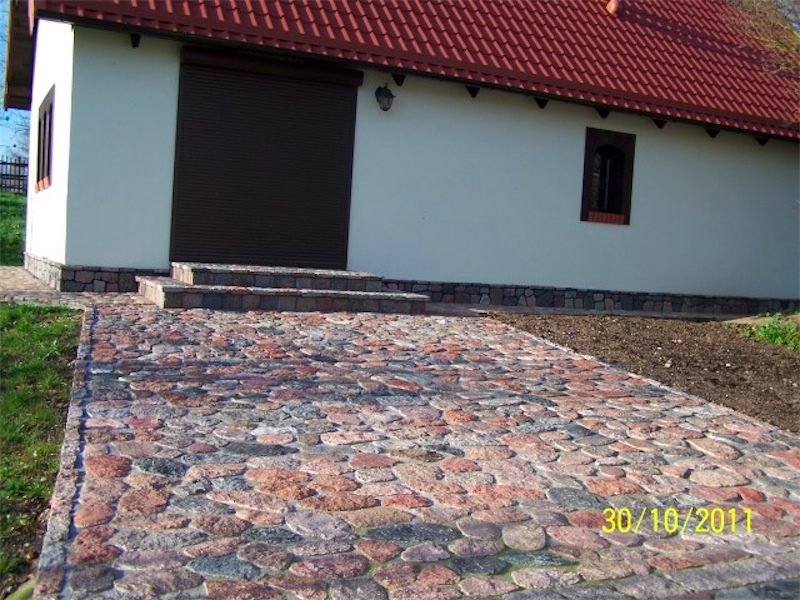 Podjazd wyłożony nieregularną kostką granitową w odcieniach czerwieni i szarości prowadzący do budynku z białymi ścianami, brązową bramą garażową i schodami z tego samego kamienia.