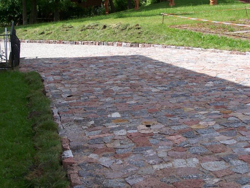 Ścieżka ułożona z nieregularnych, wielobarwnych kamieni granitowych, częściowo zacieniona, z obrzeżem z kostki granitowej i trawiastym poboczem.